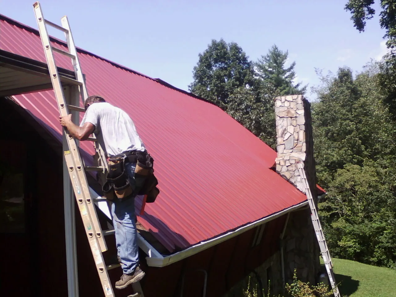 Roofer on a ladder beside a red metal roof and stone chimney — Soffit & Fascia Repair by Roofing Brothers in Williamsburg